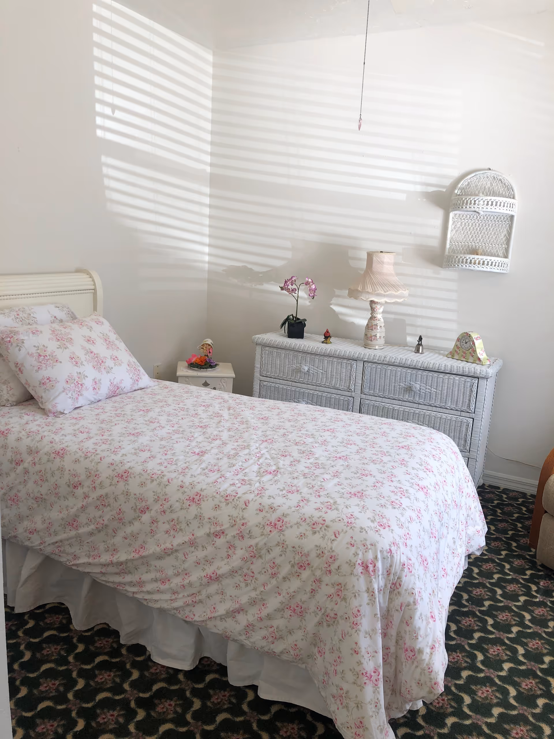 Sunlit bedroom with a floral-covered twin bed, white wicker dresser and lamp, and patterned carpet.