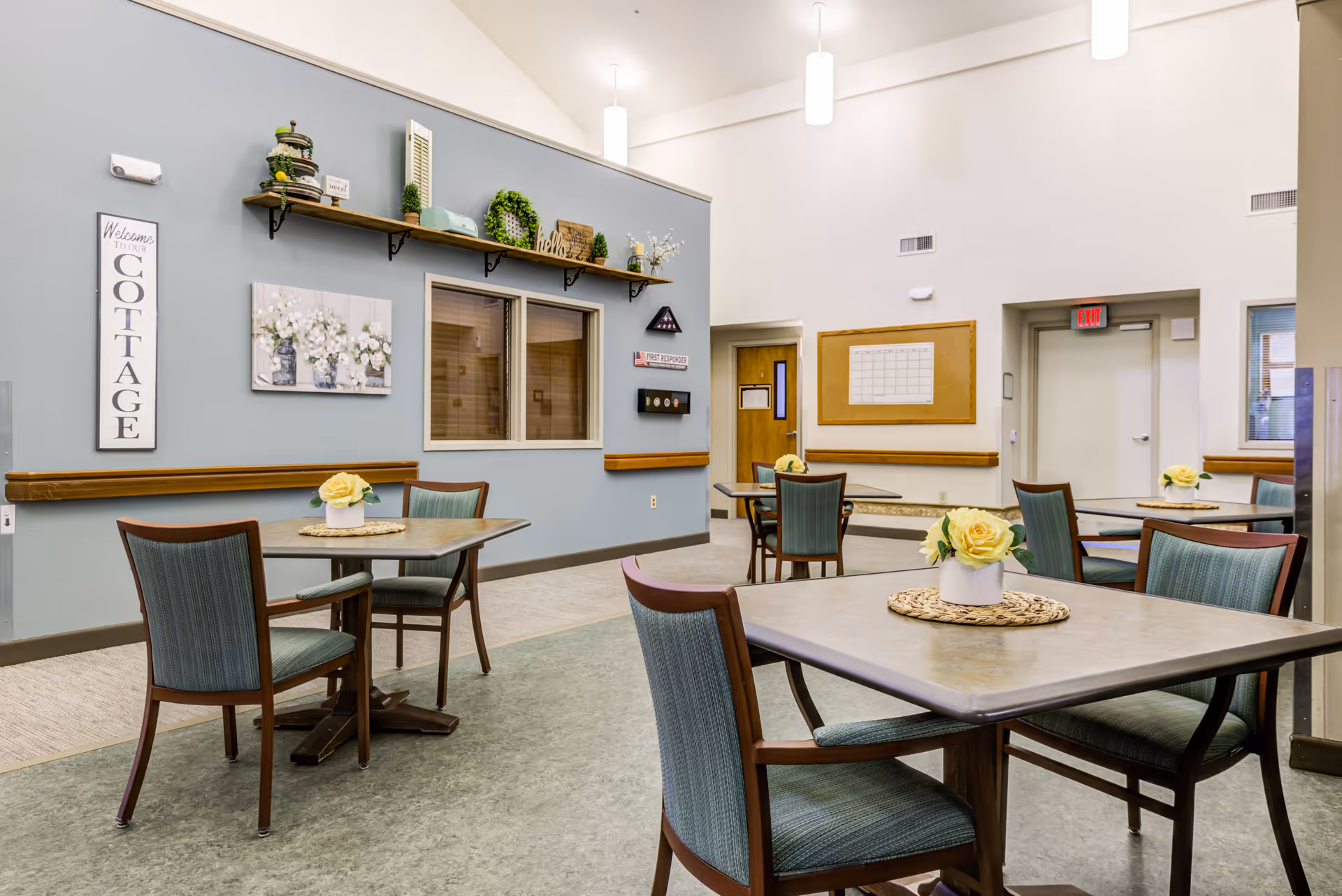 A bright and clean dining area in a senior living facility with several tables and chairs. Each table has a small floral centerpiece with yellow flowers. The walls are painted light blue and white, decorated with shelves holding plants and decorative items, a 'Welcome to our Cottage' sign, and a bulletin board. There are windows and doors visible, along with ceiling lights providing ample illumination.