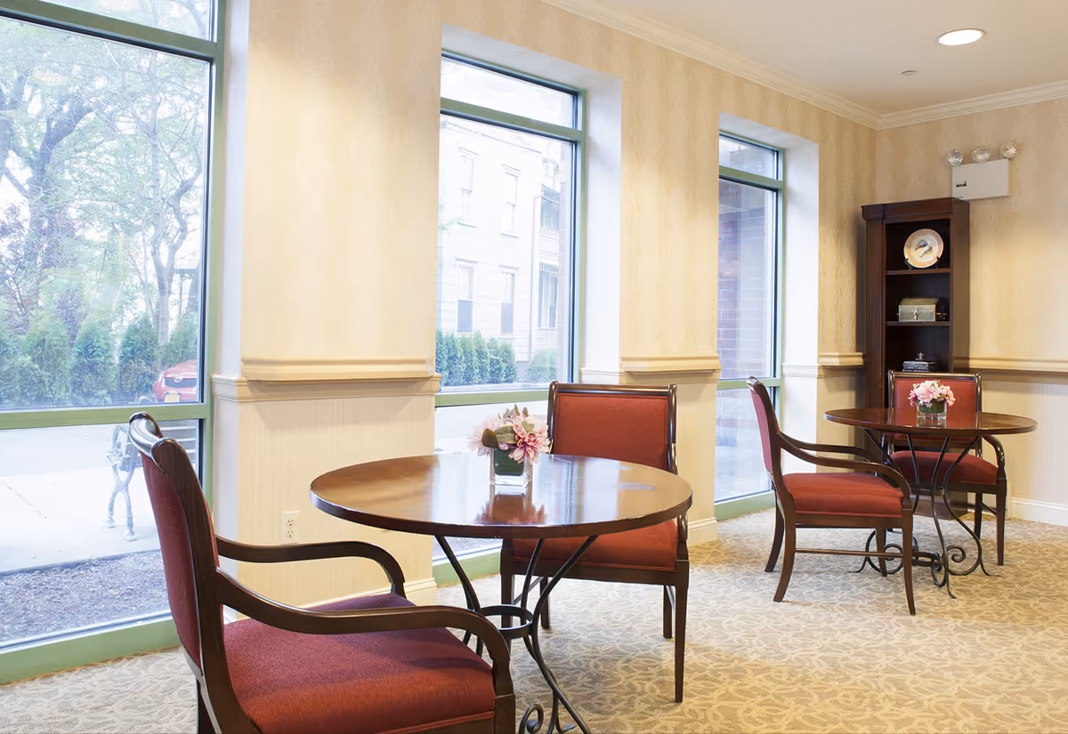 A bright and cozy seating area with two round wooden tables, each surrounded by two red upholstered chairs with wooden arms. Each table has a small floral arrangement in a glass vase. The room has large windows letting in natural light, beige patterned wallpaper, and a carpeted floor. A wooden shelving unit with decorative items is visible in the corner.