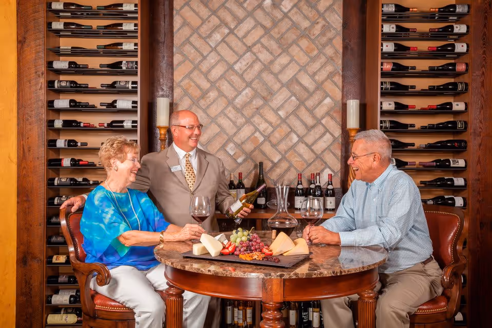 Two elderly people sitting at a round table with a man standing between them holding a wine bottle. The table has a cheese and fruit platter, wine glasses, and a wine decanter. Behind them are shelves filled with wine bottles and a decorative brick wall.