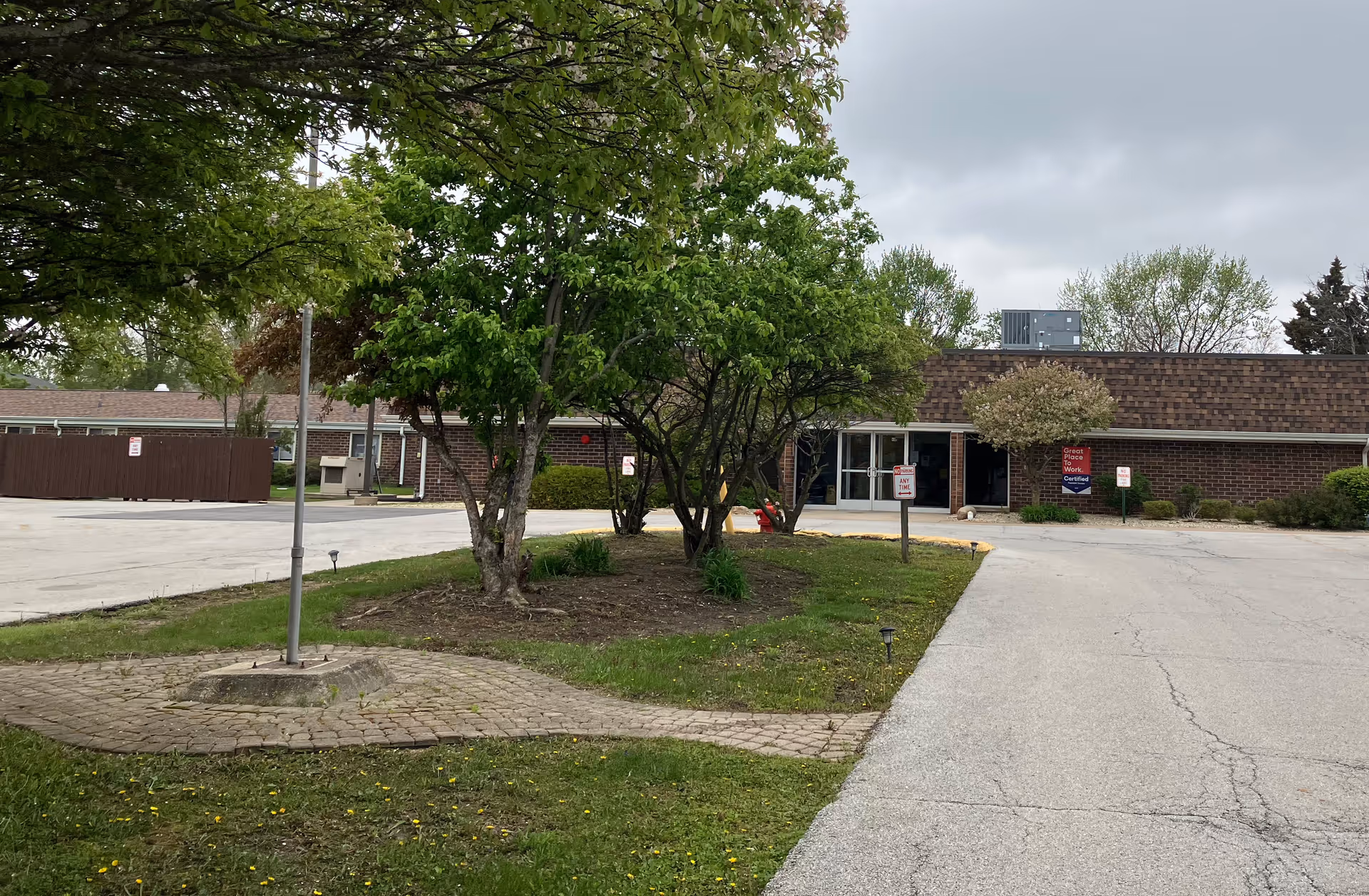 Front entrance of a single-story brick senior living facility with a driveway, trees, and a small landscaped island.