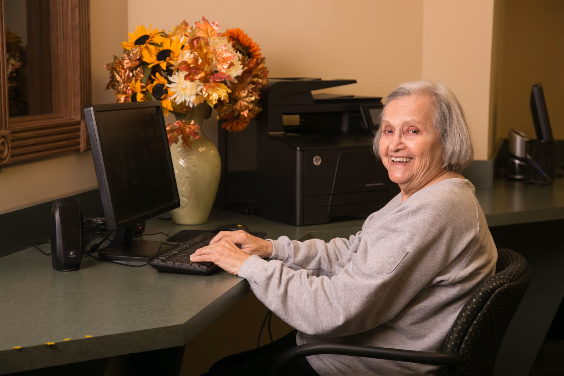 An elderly woman with gray hair is sitting at a desk using a computer keyboard. She is smiling and looking towards the camera. On the desk, there is a computer monitor, a speaker, a printer, and a vase with colorful flowers. The setting appears to be a cozy indoor area with warm lighting.
