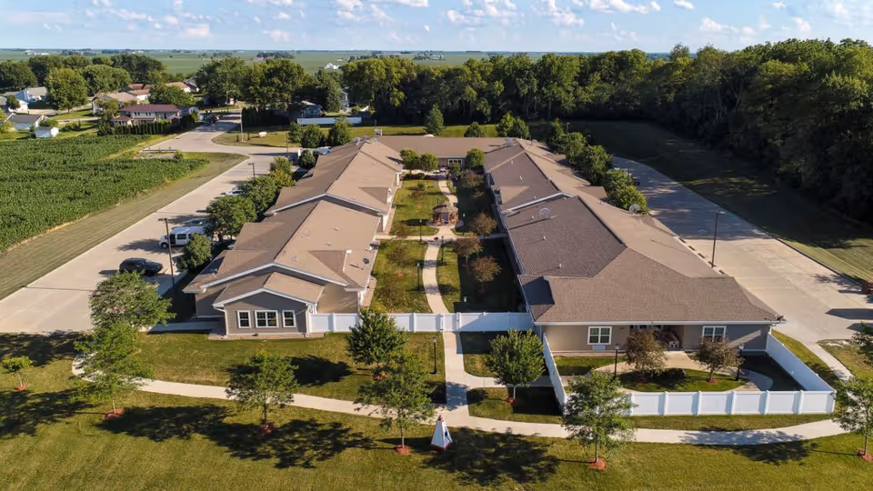 Aerial view of Homestead Assisted Living & Memory Care of Morrison showing two long single-story buildings with beige roofs and light-colored walls, separated by a landscaped courtyard with a walking path, trees, and shrubs. The facility is surrounded by green lawns, trees, and a white fence, with a road and farmland visible in the background under a partly cloudy sky.