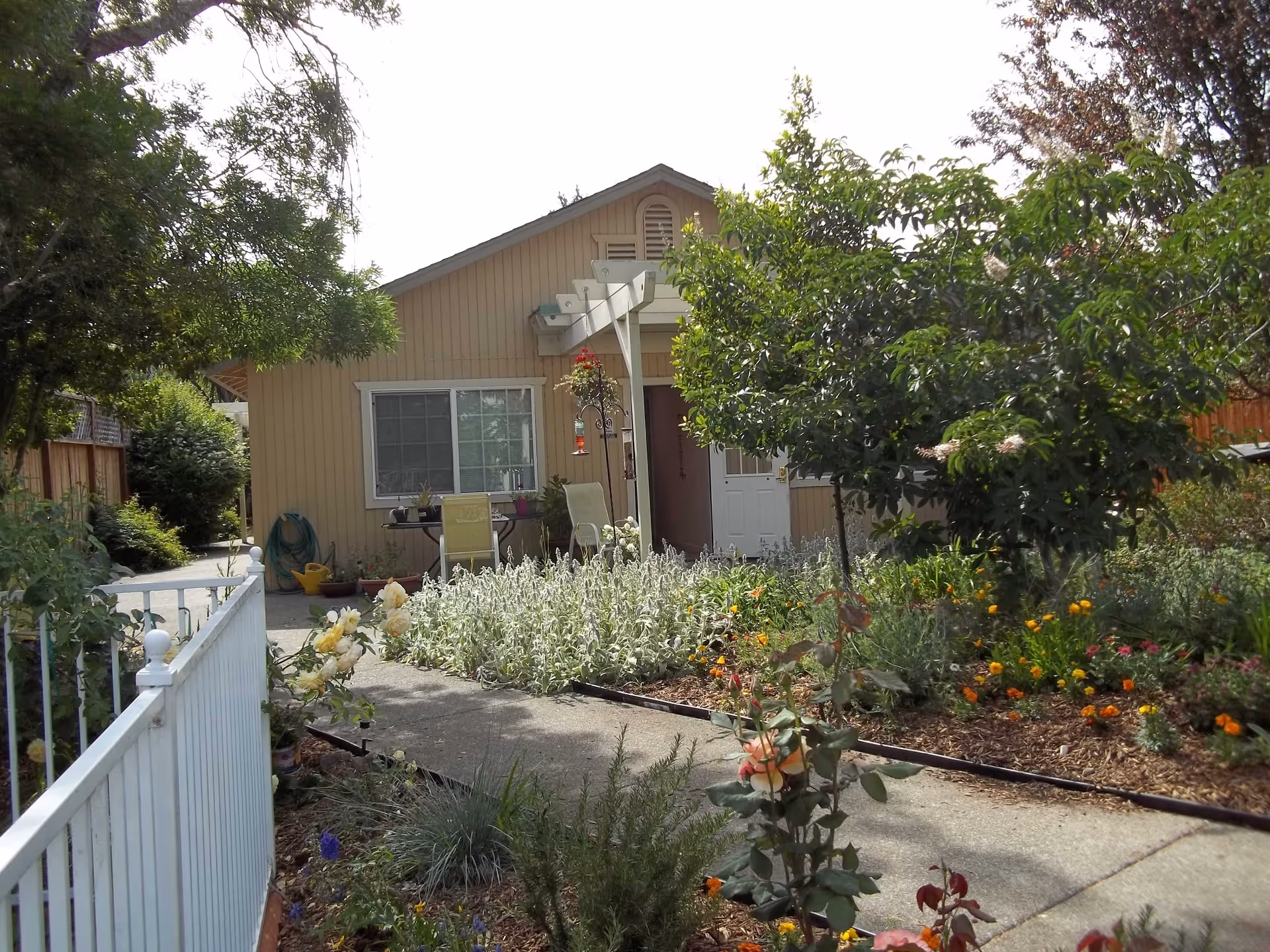 A single-story beige house with a white door and a window, surrounded by a garden with various plants and flowers. There is a concrete pathway leading to the entrance, a white fence on the left side, and two chairs with a small table near the window. Trees and shrubs are visible around the house.
