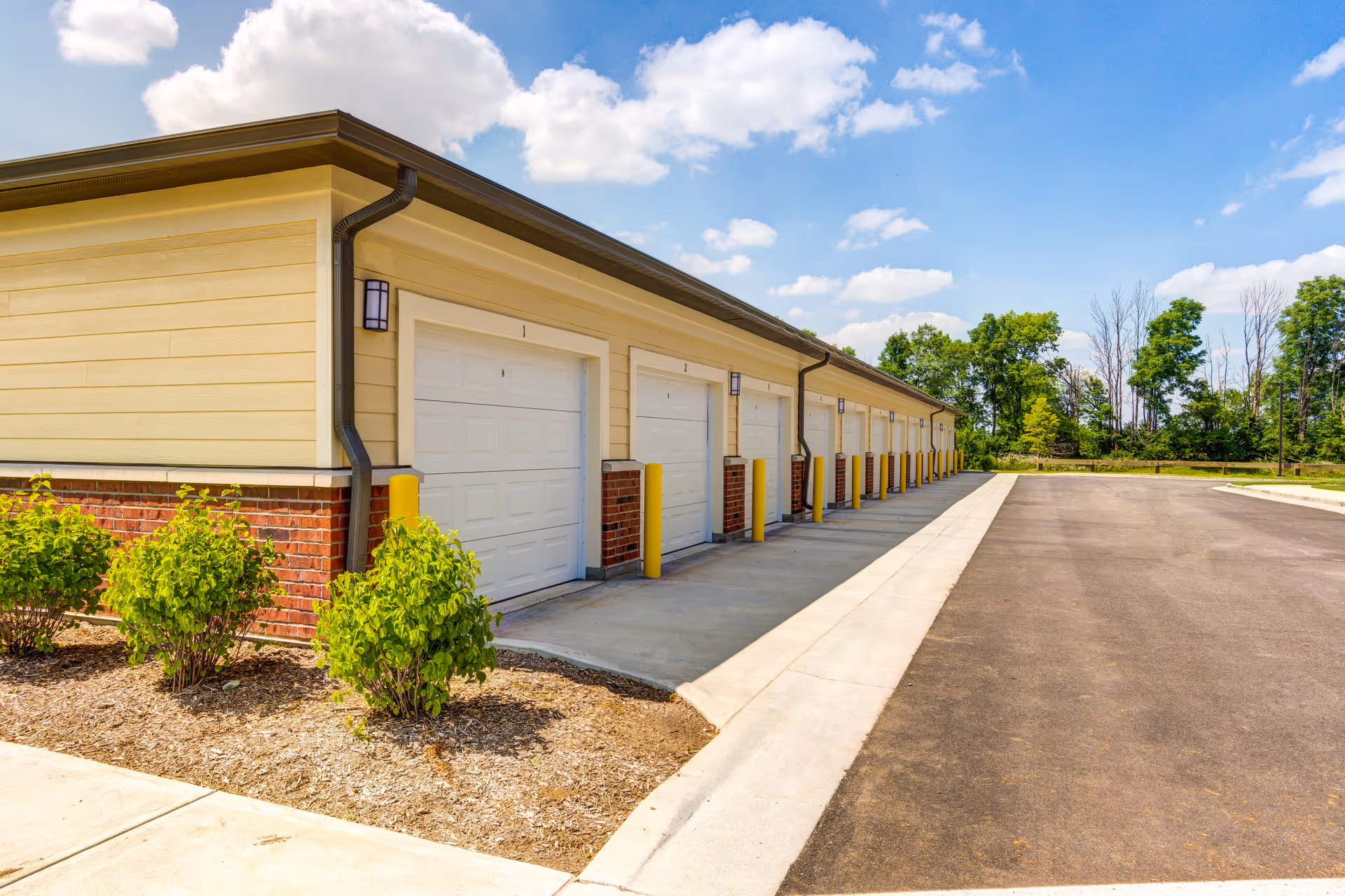 A row of closed white garage doors on a single-story building with beige siding and red brick accents, situated next to a paved driveway and landscaped with small green bushes under a partly cloudy blue sky.