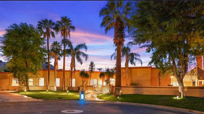 Exterior view of a senior living facility at dusk with palm trees and other greenery in front of a single-story building under a colorful sky.