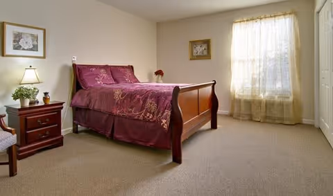 A bedroom with a wooden bed frame and a maroon bedspread. There is a nightstand with a lamp and a small plant on it to the left of the bed. The room has beige walls, a window with sheer curtains, and two framed pictures on the walls.