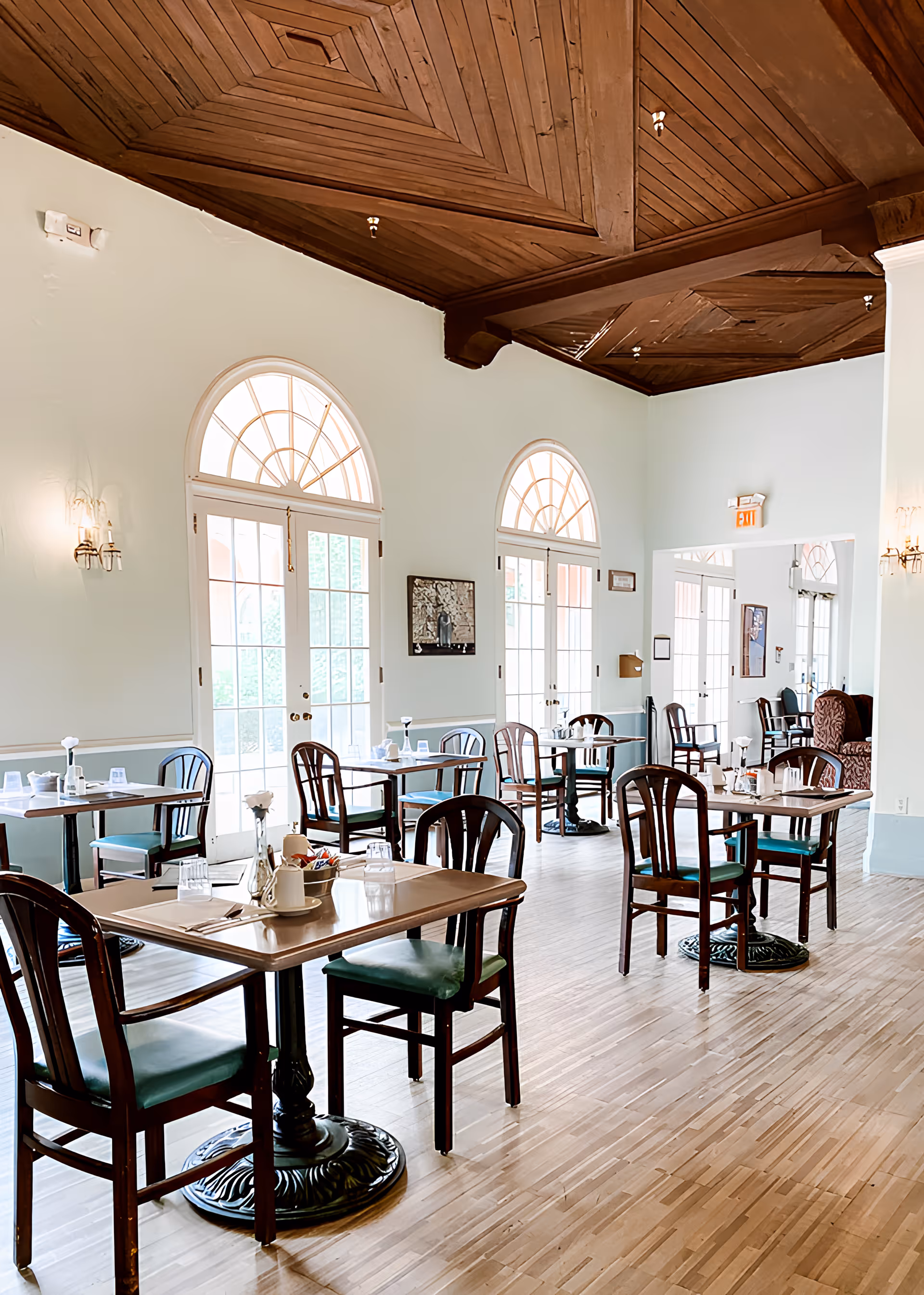 Bright dining room with a decorative wooden ceiling, arched windows, and several set tables and chairs on a light wood floor.