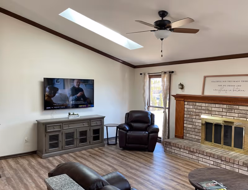 Bright living room with a wall-mounted TV above a gray media console, a leather recliner, skylight, and a brick fireplace.