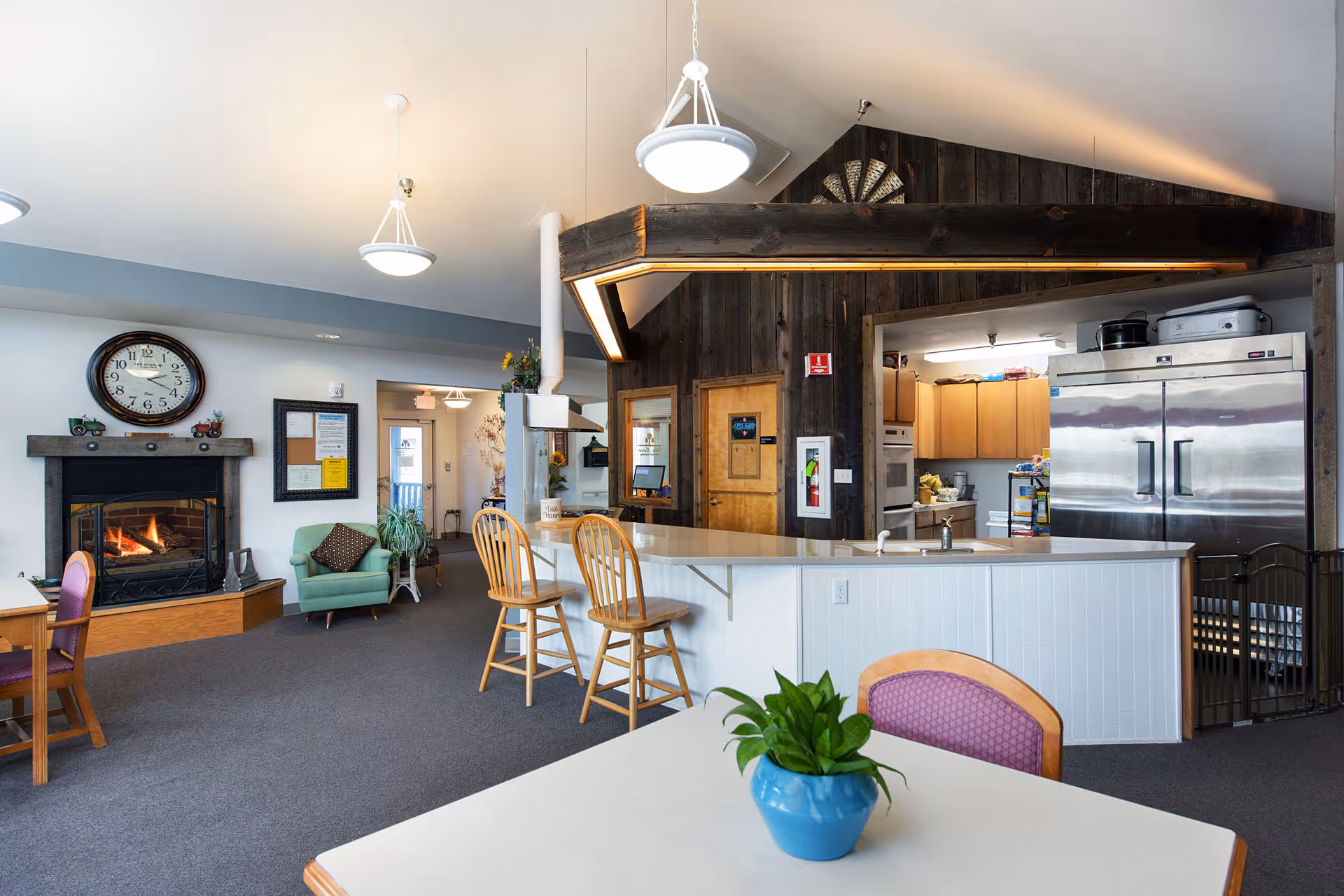 Interior view of a senior living facility common area featuring a kitchen with a large stainless steel refrigerator, wooden cabinets, and a counter with two wooden bar stools. There is a cozy fireplace with a clock above it, a green armchair, and a table with a purple chair and a blue pot with a green plant. The space has a warm and inviting atmosphere with soft lighting and a mix of wood and modern elements.