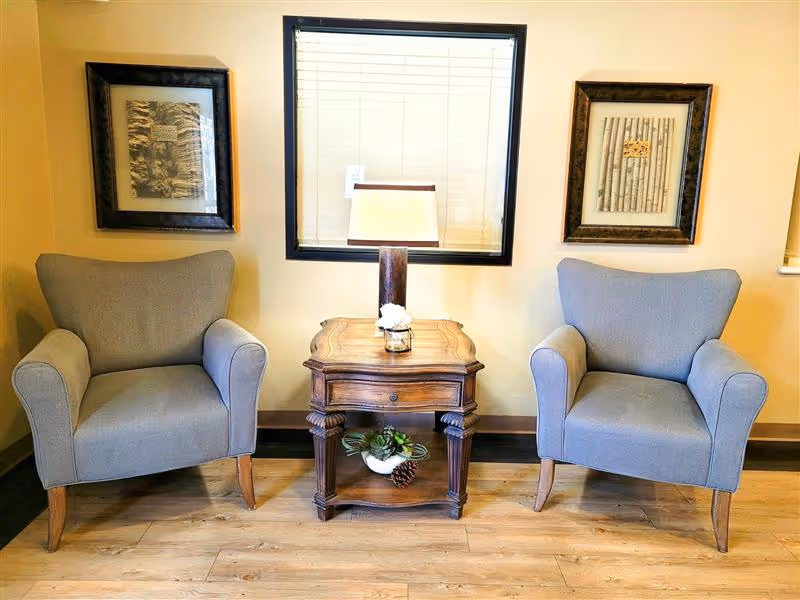 Two gray upholstered armchairs with wooden legs are positioned on either side of a wooden side table with a lamp and a small decorative plant. Behind the table is a window with closed blinds, and on the wall above each chair hangs framed artwork. The room has light-colored walls and wooden flooring.