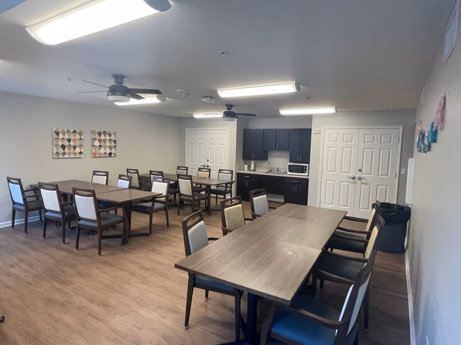 A communal dining room with several wooden tables and chairs arranged neatly. The room has light-colored walls, two ceiling fans, and overhead fluorescent lights. There is a small kitchenette area with dark cabinets, a microwave, and a sink. Two sets of double doors are visible on the far wall, and there are two geometric patterned artworks hanging on one wall.