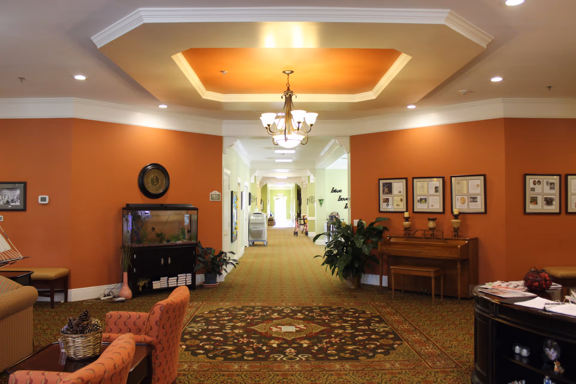 Interior view of a senior living facility hallway with orange walls, patterned carpet, a chandelier, framed pictures on the walls, a fish tank, plants, a piano, and seating areas.