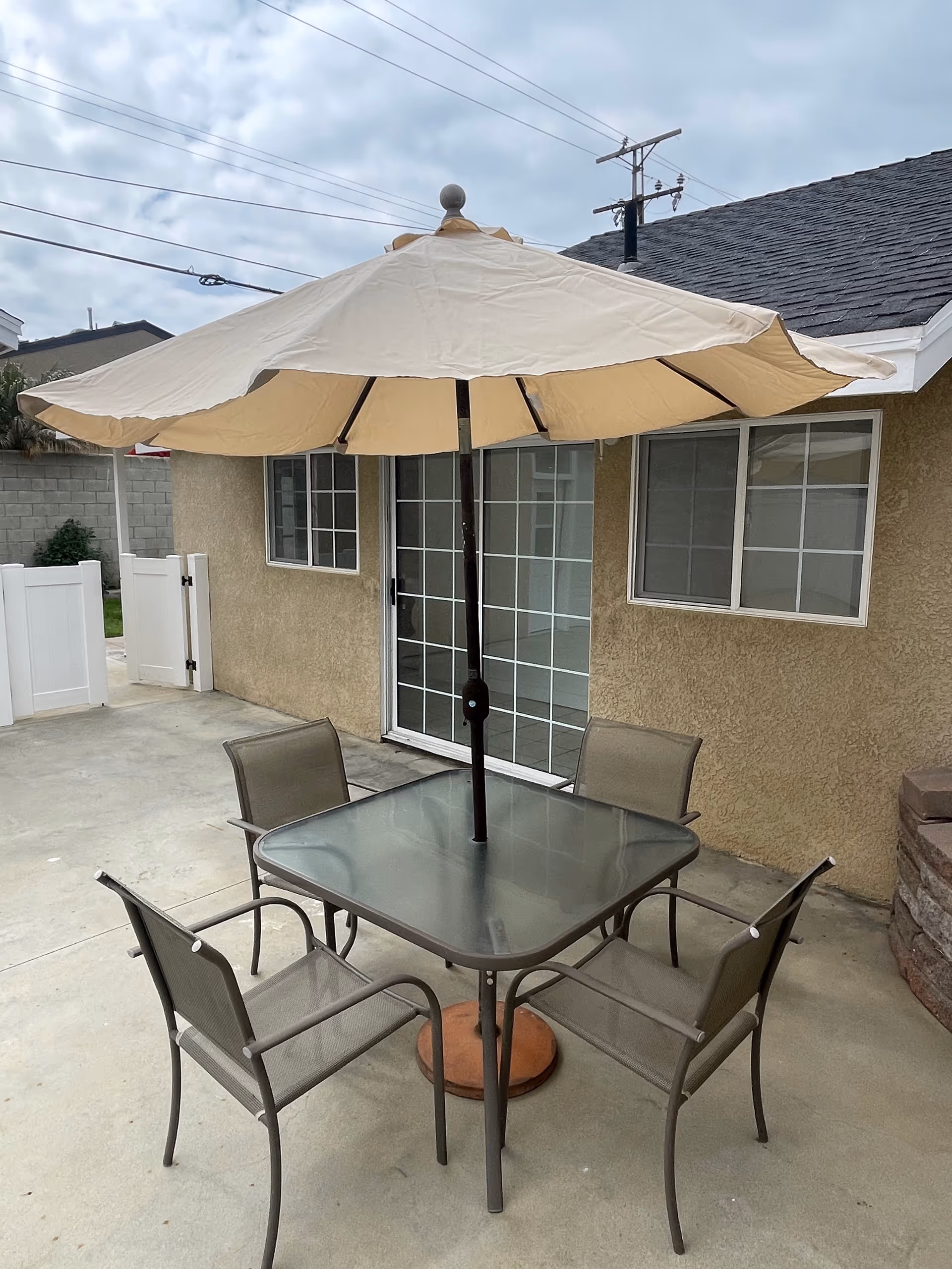 Outdoor patio area with a square glass table and four metal chairs with mesh seats and backs. A large beige patio umbrella is centered in the table. The patio is adjacent to a beige stucco building with sliding glass doors and windows. The ground is concrete and there is a white fence and gate in the background.