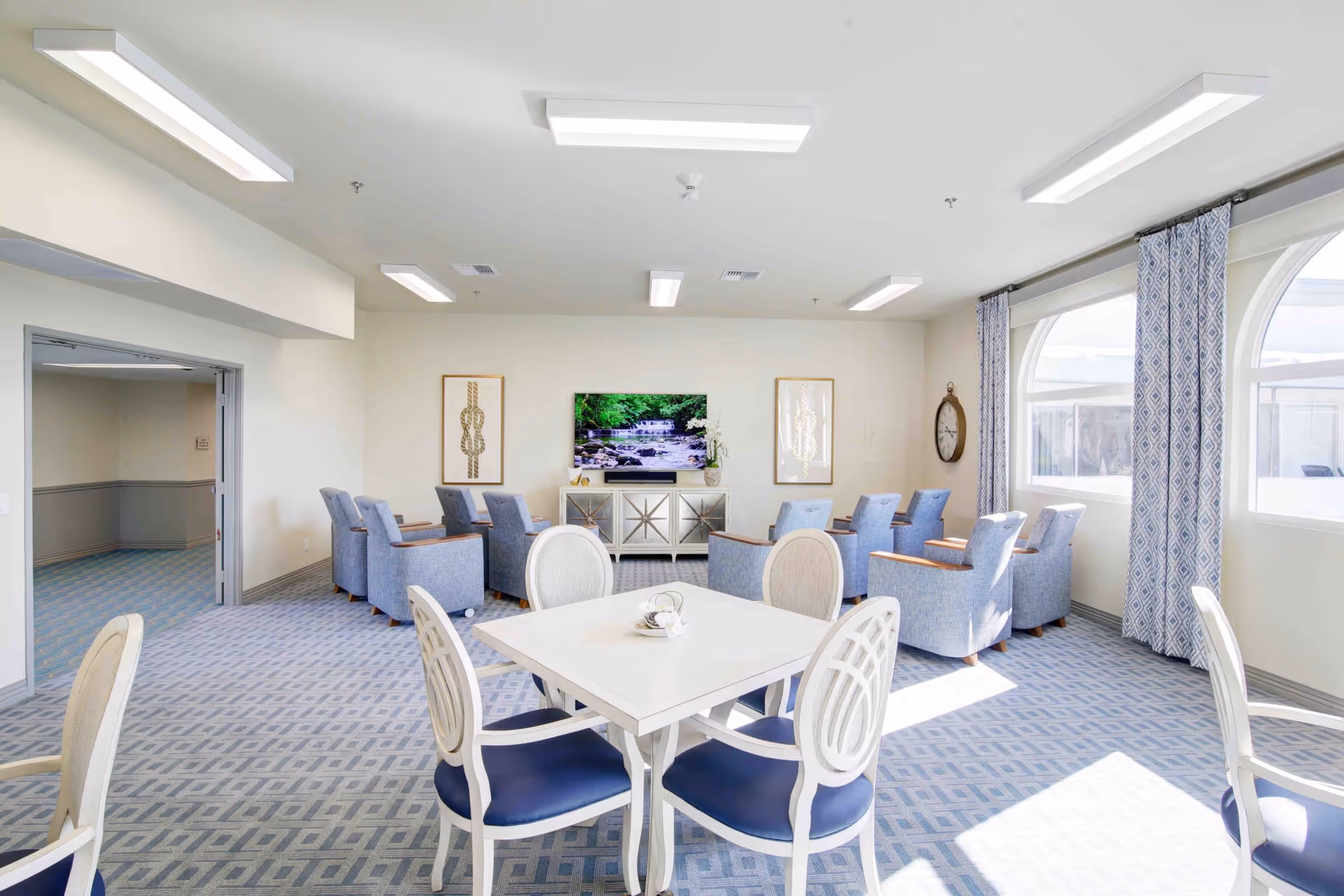A bright and spacious senior living common area with a white table and four chairs in the foreground, and a row of blue armchairs facing a wall-mounted TV displaying a nature scene. The room has large windows with patterned curtains allowing natural light to fill the space, light-colored walls, and a patterned carpet.