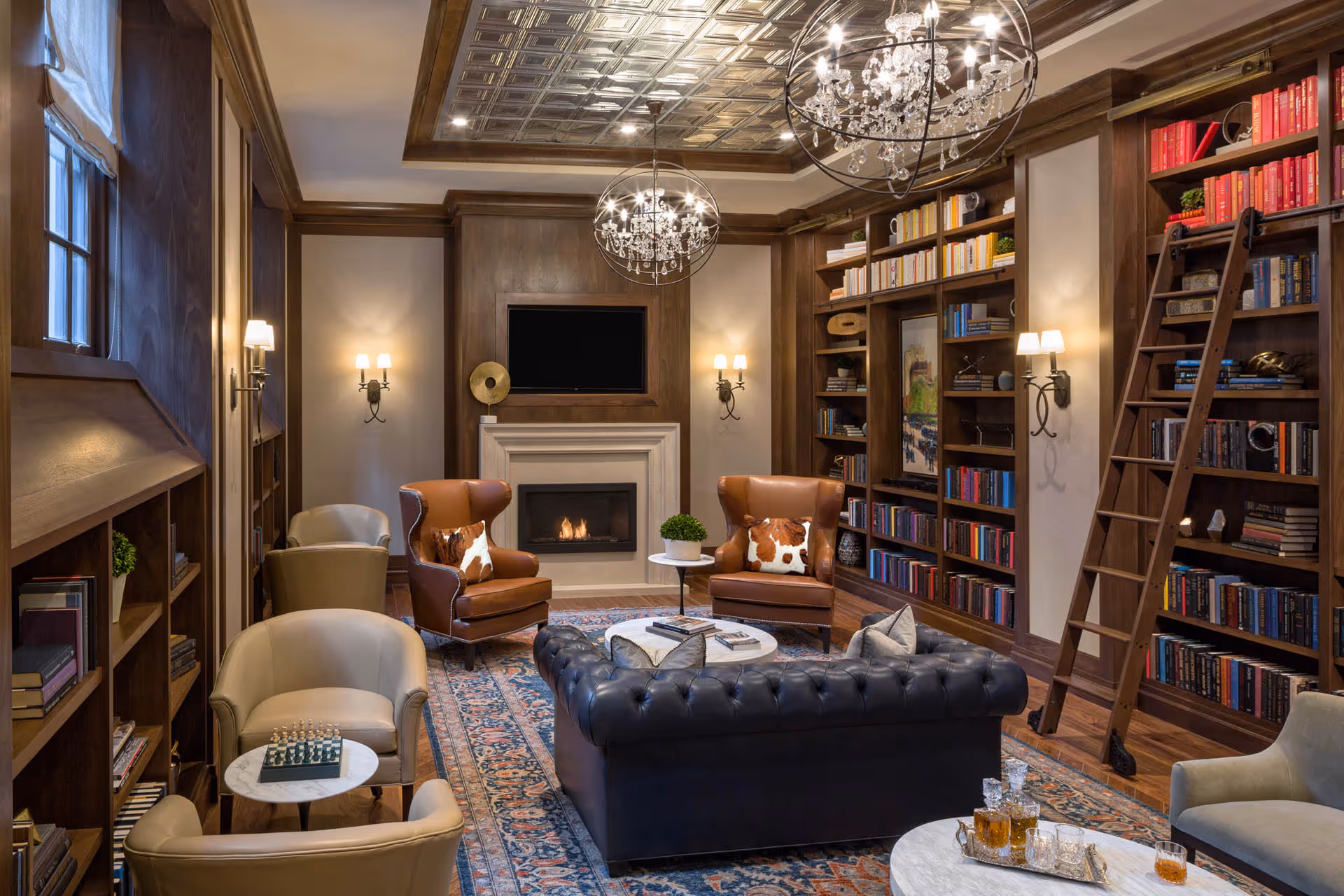 Cozy wood-paneled library lounge with built-in bookshelves, leather seating, a fireplace, chandeliers, and a rolling ladder.