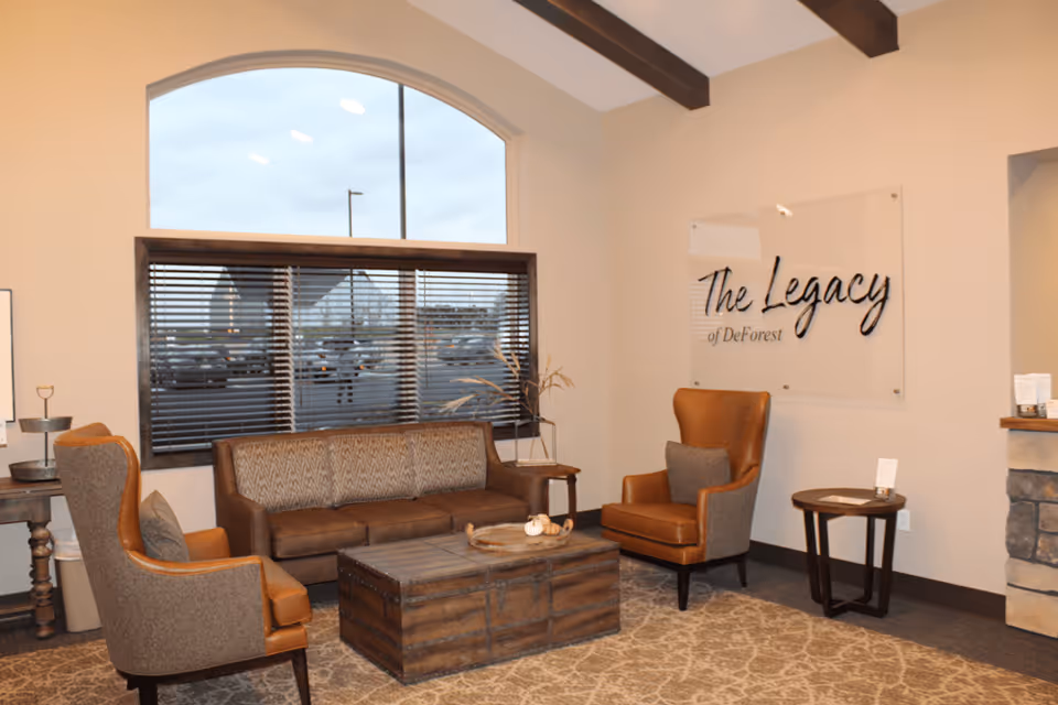 A cozy seating area inside The Legacy of DeForest facility featuring a brown leather sofa, two brown armchairs with gray cushions, a wooden coffee table, side tables, a large window with blinds, and a wall sign that reads 'The Legacy of DeForest'.