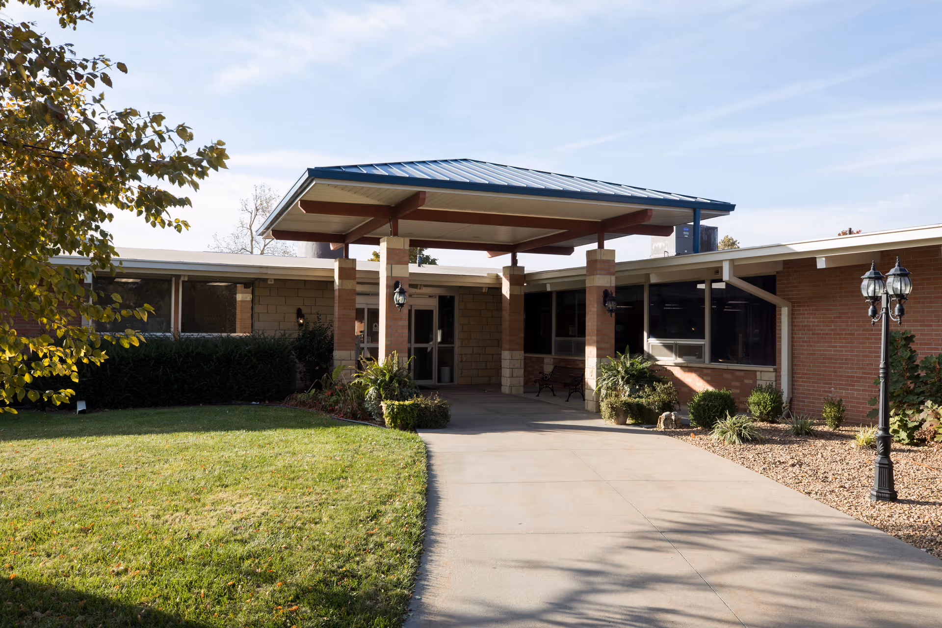 Exterior front entrance of a single-story brick building with a covered canopy, walkway, lawn, and lamppost.