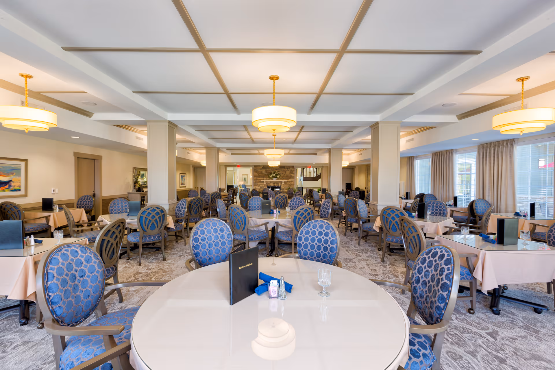A spacious dining room with multiple round and rectangular tables covered with light-colored tablecloths. Each table is surrounded by blue upholstered chairs with a circular pattern. The room features a coffered ceiling with hanging light fixtures, large windows with curtains allowing natural light, and a carpeted floor with a subtle pattern. There are menus and glassware on the tables, and the overall ambiance is bright and inviting.