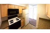 Small kitchen with wooden cabinets, electric range and microwave, refrigerator, and granite countertops leading toward a doorway.