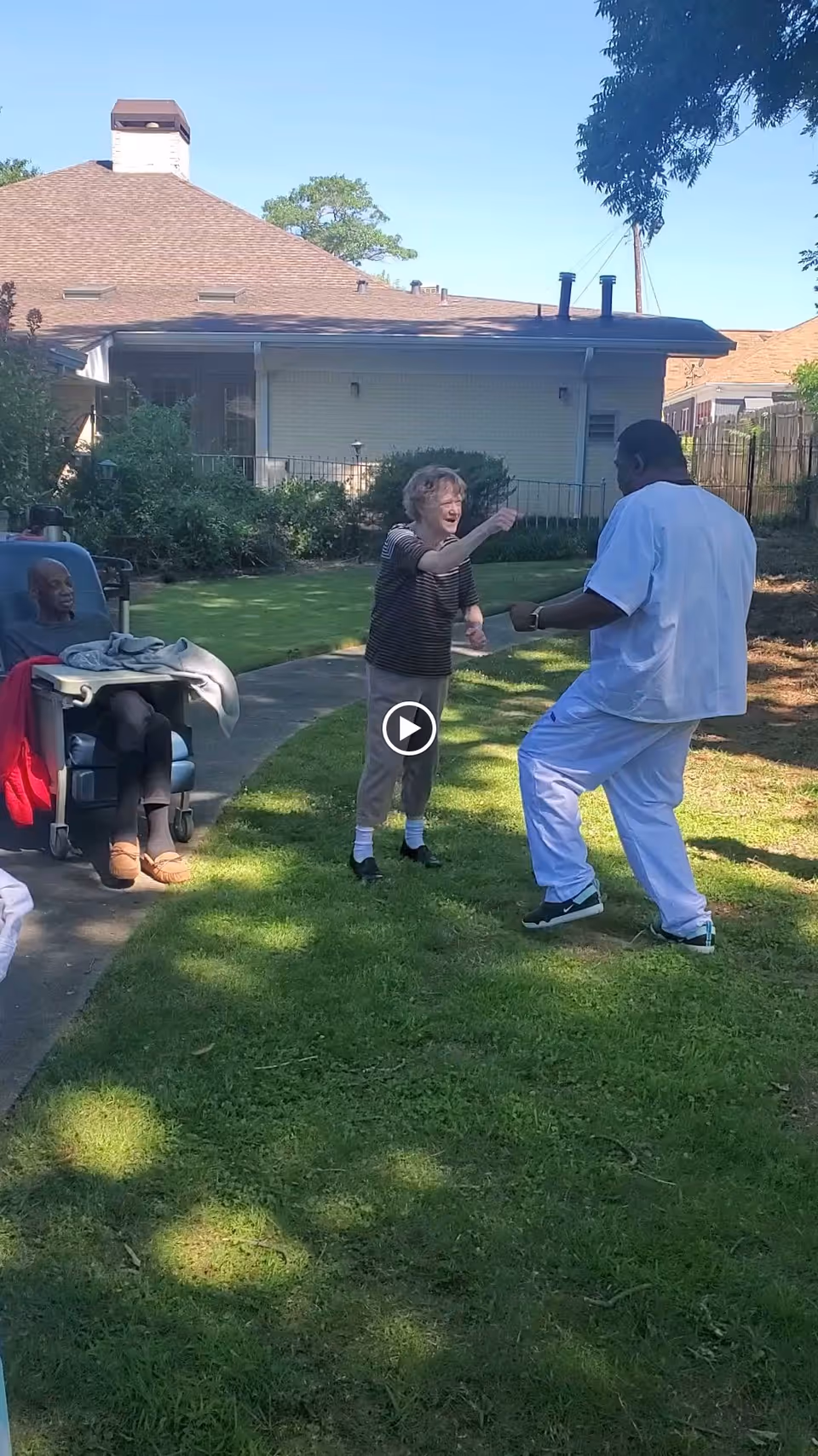 An elderly woman reaches toward a caregiver in a grassy yard while another resident sits in a wheelchair near a residential building.