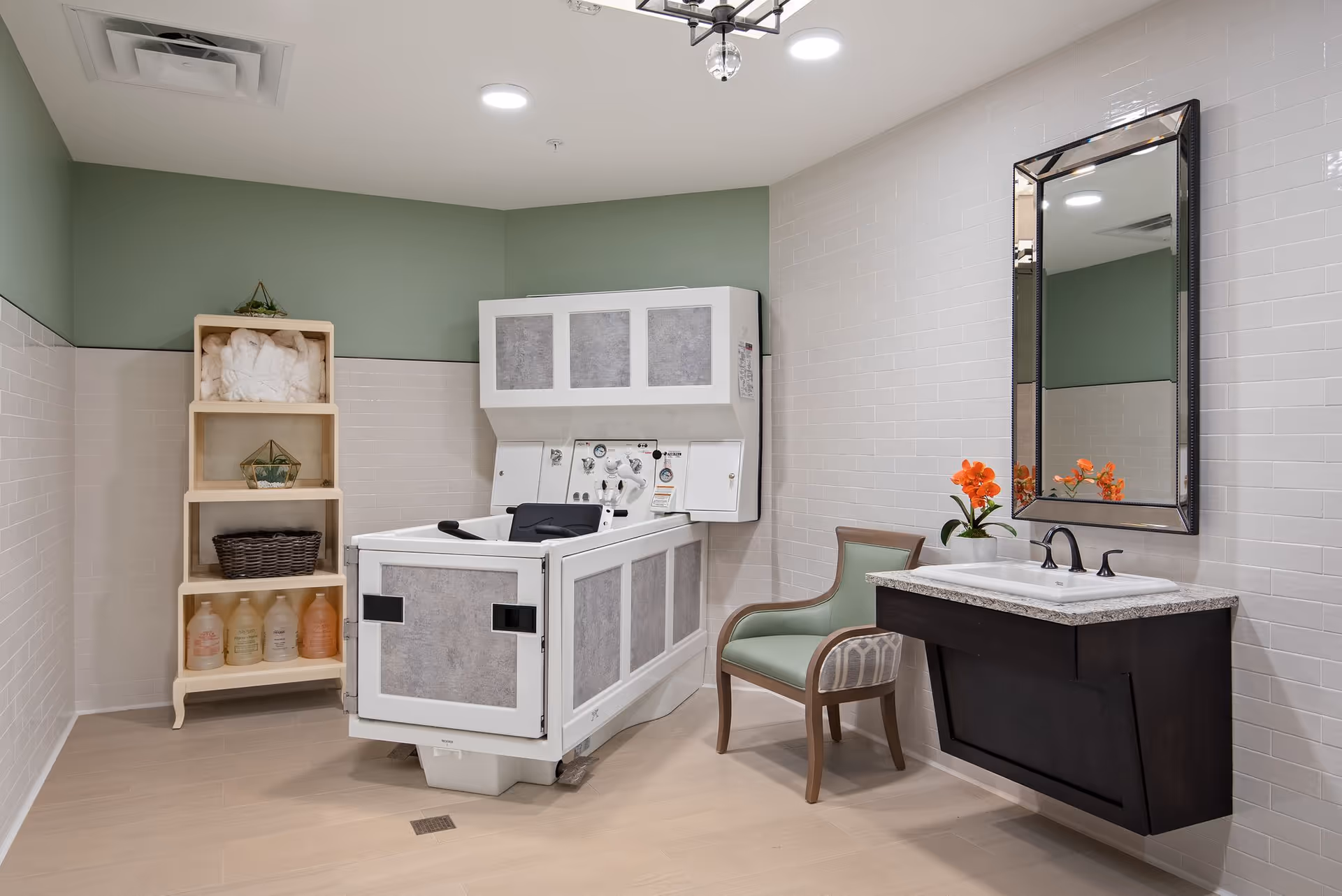 A clean and modern bathing room with a specialized bathtub for assisted bathing, a wooden shelving unit holding towels and bottles, a green upholstered chair, a sink with a granite countertop, a large mirror, and a small vase with orange flowers.
