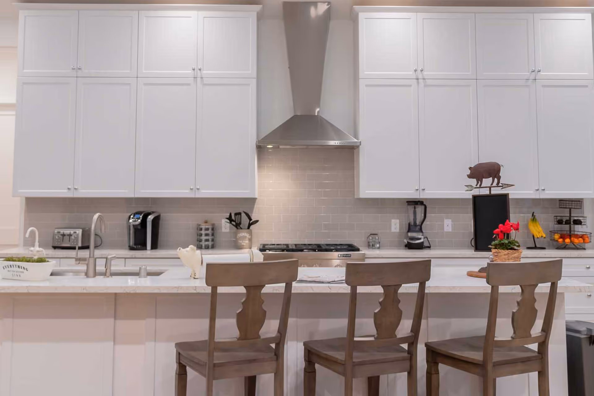 Modern kitchen with white cabinets, a stainless steel range hood, a gas stove, and a white marble countertop with three wooden chairs. The countertop has a sink, a coffee maker, a toaster, kitchen utensils, a blender, a fruit basket with bananas and oranges, and a small potted plant with red flowers.