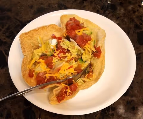 A taco salad served in a crispy tortilla bowl on a white plate, topped with shredded cheddar cheese, lettuce, diced tomatoes, and a dollop of sour cream, with a fork resting on the plate.