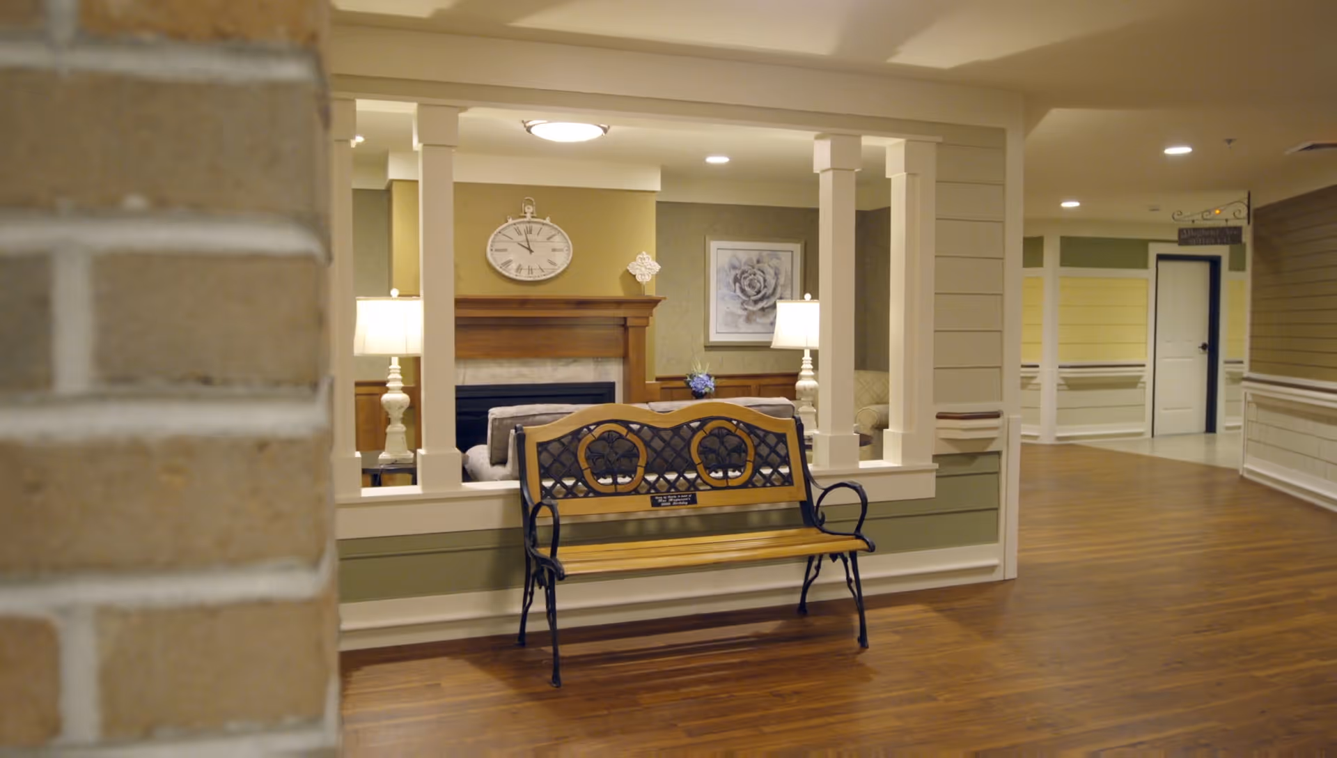 Interior view of a senior living facility hallway with wooden flooring and a decorative bench in the foreground. Through a large window opening, a cozy living room area is visible with a fireplace, a clock above it, two lamps, and a framed flower picture on the wall.