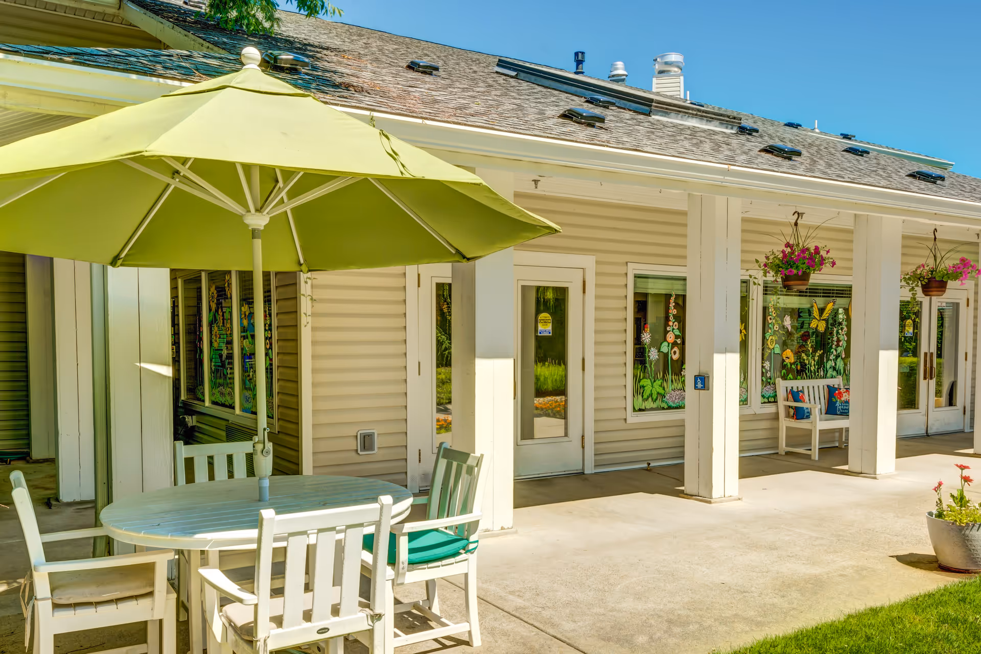 Outdoor patio area at The Suites Assisted Living and Memory Care featuring a round white table with four white chairs, one with a green cushion, and a large green umbrella providing shade. The building exterior has beige siding with white columns and doors, decorated with colorful flower and butterfly window art and hanging flower pots.