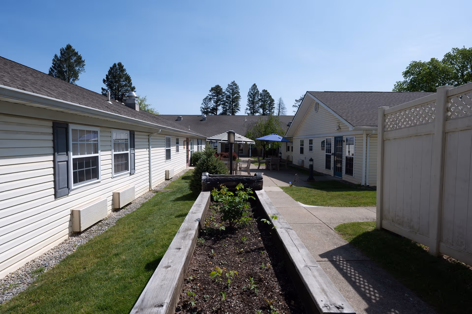 Outdoor courtyard area between two single-story buildings with beige siding and gray shutters. There is a raised garden bed with plants in the center, a paved walkway, green grass, patio tables with umbrellas, and trees in the background under a clear blue sky.