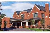 Brick senior living building with a covered porte-cochère entrance, columns, landscaped lawn, and blue sky.