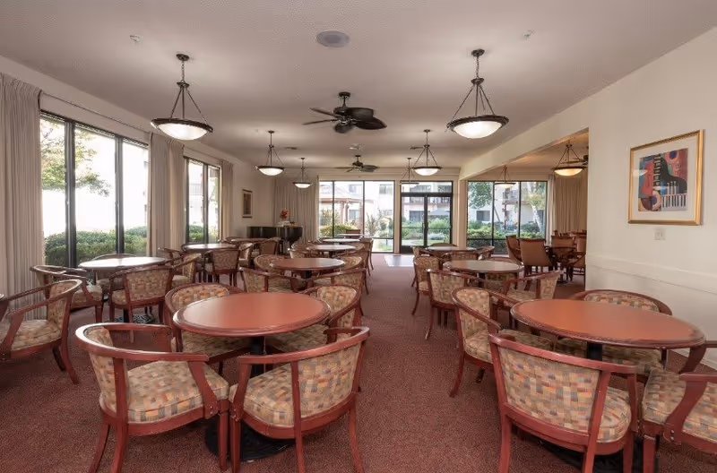 Spacious senior living dining room with multiple round tables and upholstered chairs facing large windows and glass doors to an outdoor courtyard.