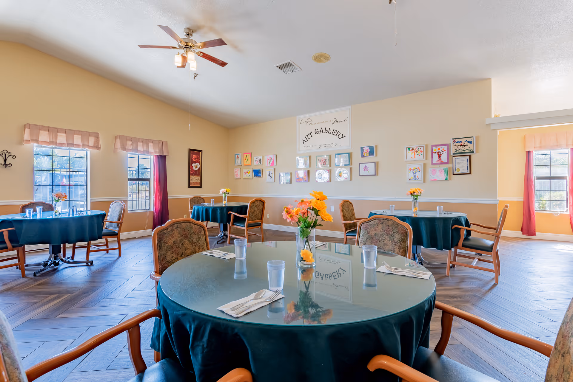 Bright dining room with round tables covered in green tablecloths, floral centerpieces, chairs, windows, and an 'Art Gallery' display on the far wall.