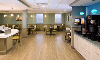 A spacious dining area with multiple round tables and chairs arranged neatly. The room features wood flooring, green walls, and several windows letting in natural light. On the right side, there is a beverage station with coffee machines and a soda dispenser under hanging blue pendant lights.