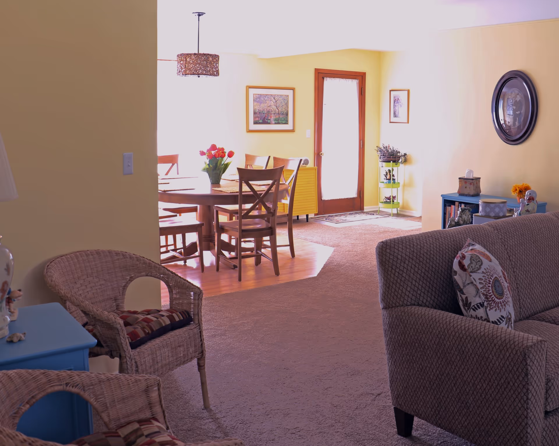 Interior view of a senior care facility showing a cozy living room area with a patterned sofa and wicker chairs, leading into a dining area with a wooden table and chairs. The walls are painted yellow and decorated with framed pictures. A door with a sheer curtain lets in natural light.