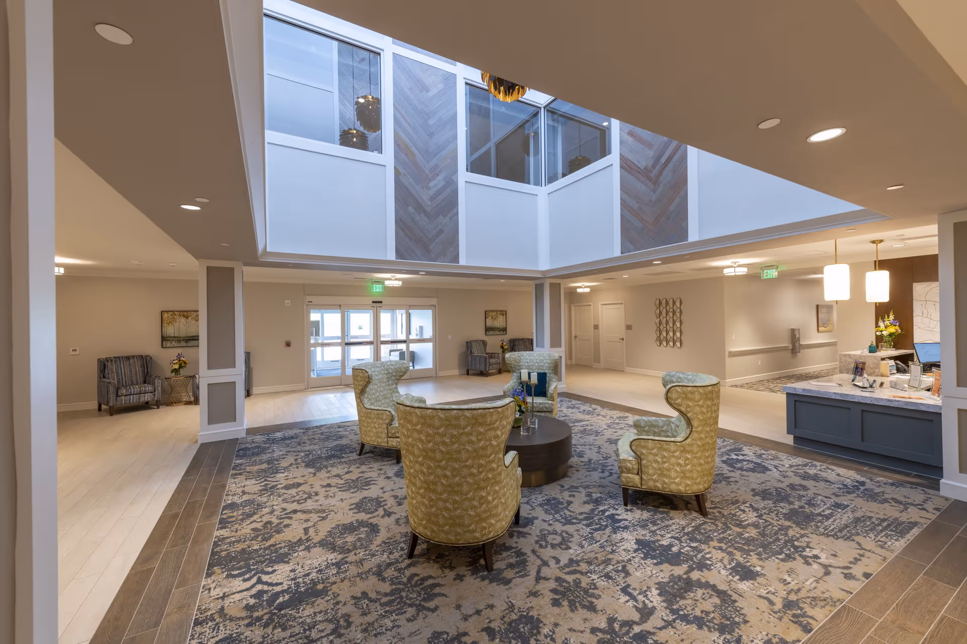 Spacious senior living facility lobby with a large patterned rug, four green upholstered armchairs arranged around a round wooden coffee table, and a reception desk with pendant lights on the right. The area features a high ceiling with a skylight and wood panel accents, light-colored walls, and multiple seating areas with armchairs along the walls.