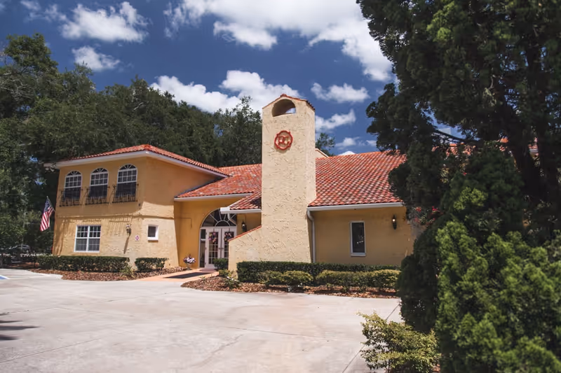 Exterior view of a yellow stucco building with a red tile roof under a blue sky with scattered clouds. The building has multiple windows, an American flag on the left side, and is surrounded by greenery and trees.