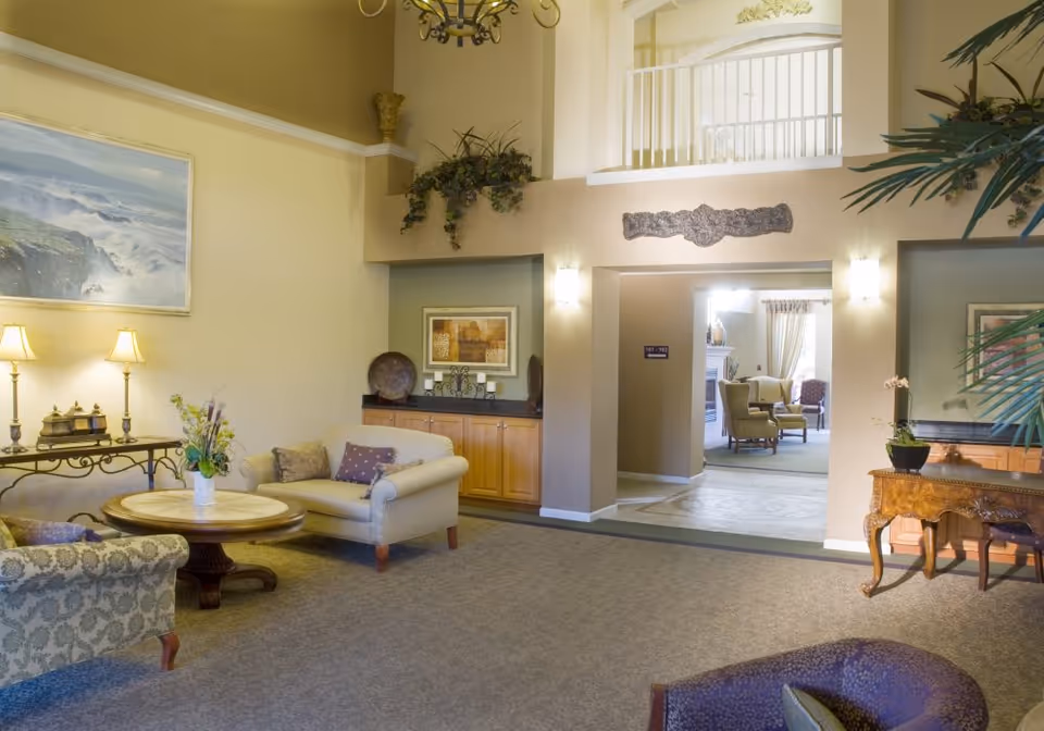 A spacious and warmly lit senior living facility common area with comfortable armchairs and sofas arranged around a wooden coffee table. The room features soft beige walls, decorative plants, framed artwork, and a chandelier hanging from the ceiling. In the background, another seating area with armchairs and a fireplace is visible through an open doorway.