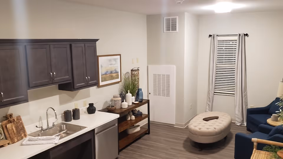 A cozy interior room featuring a small kitchen area with dark cabinets, a stainless steel sink, and a dishwasher. Next to the kitchen is a wooden shelf decorated with vases and plants. The room also includes a seating area with a blue armchair, a round beige ottoman, and a window with closed blinds and light gray curtains. The floor is wood-style laminate, and the walls are painted light gray.