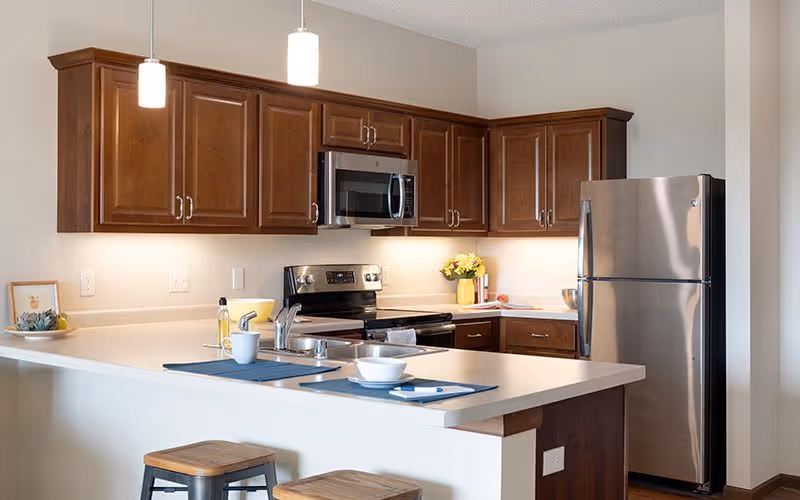 Modern kitchen with wooden cabinets, stainless steel refrigerator, stove, and microwave. The kitchen island has a sink, two stools, and place settings with a cup and bowl. Pendant lights hang above the island, and there is a small plant and a vase with flowers on the countertop.