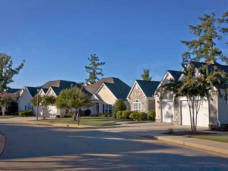 A quiet residential street in a senior living community with single-story houses featuring garages, well-maintained lawns, trees, and clear blue sky.