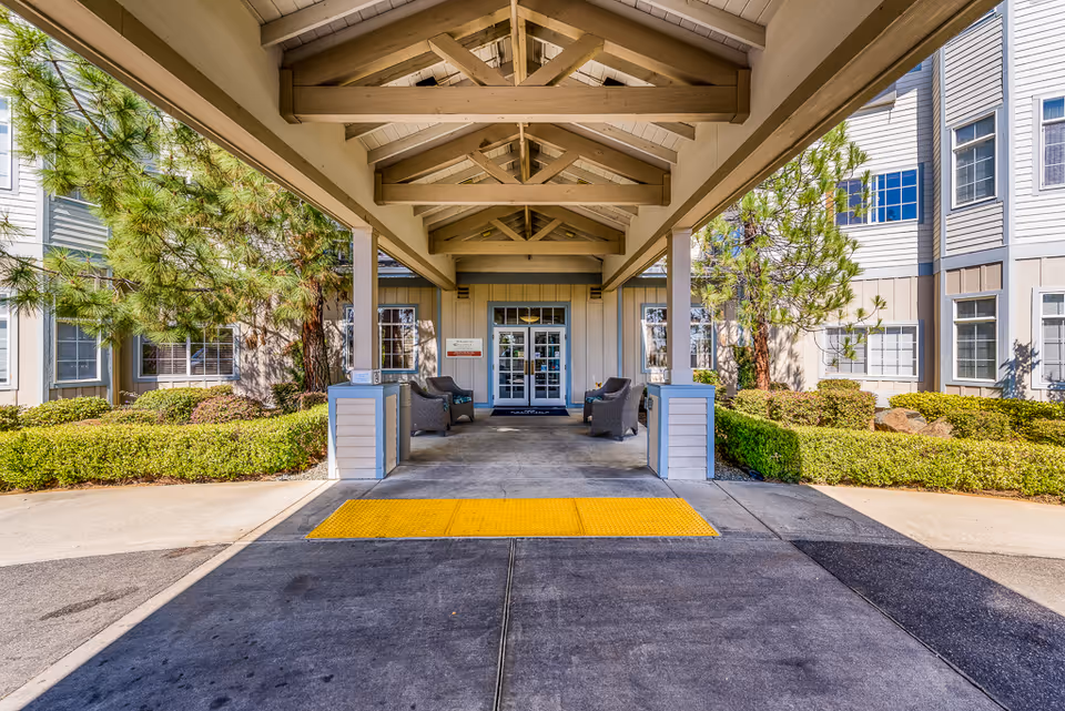 Covered entrance and porte-cochère of a senior living building with seating, landscaped bushes, and a yellow tactile pad leading to glass doors.