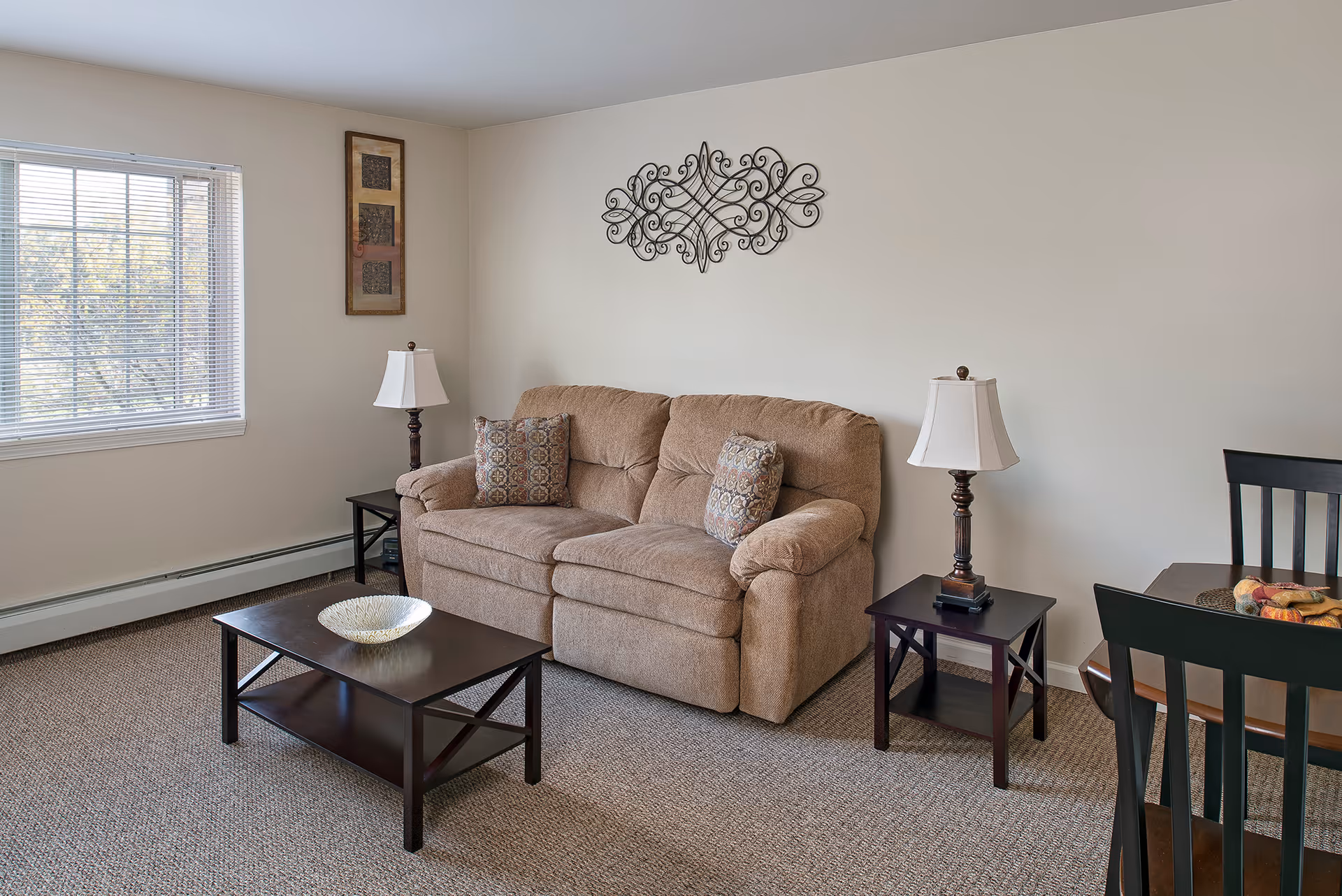 Living room with a beige loveseat, coffee table, two side tables with lamps, wall decor, window, and a small dining table partially visible.