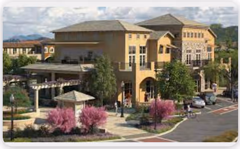 Exterior view of a multi-story senior living facility building with beige and stone facade, surrounded by landscaped gardens with pink flowering trees, a pergola, and a parking area with cars. Mountains are visible in the background under a partly cloudy sky.