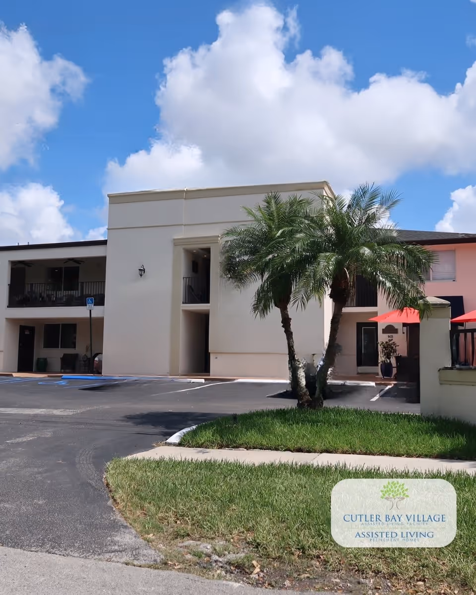 Exterior view of a two-story assisted living facility building with a beige facade, palm trees, a parking lot with handicap parking spaces, and a partly cloudy blue sky.