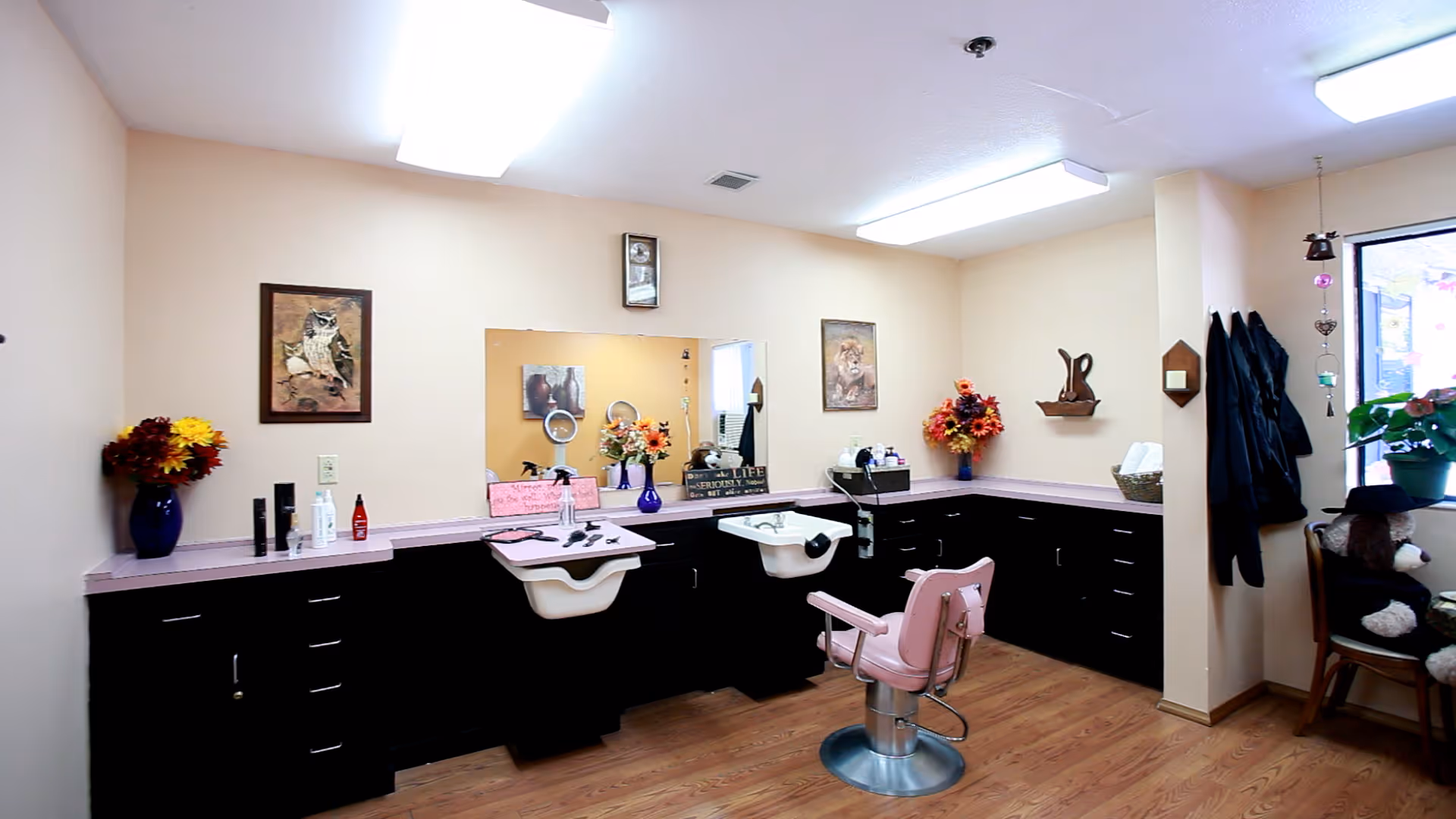 Interior of a hair salon area in a senior living facility with two white wash basins, a pink salon chair, black cabinets with pink countertops, floral decorations, framed pictures on the walls, and a window with plants and a teddy bear on a chair nearby.