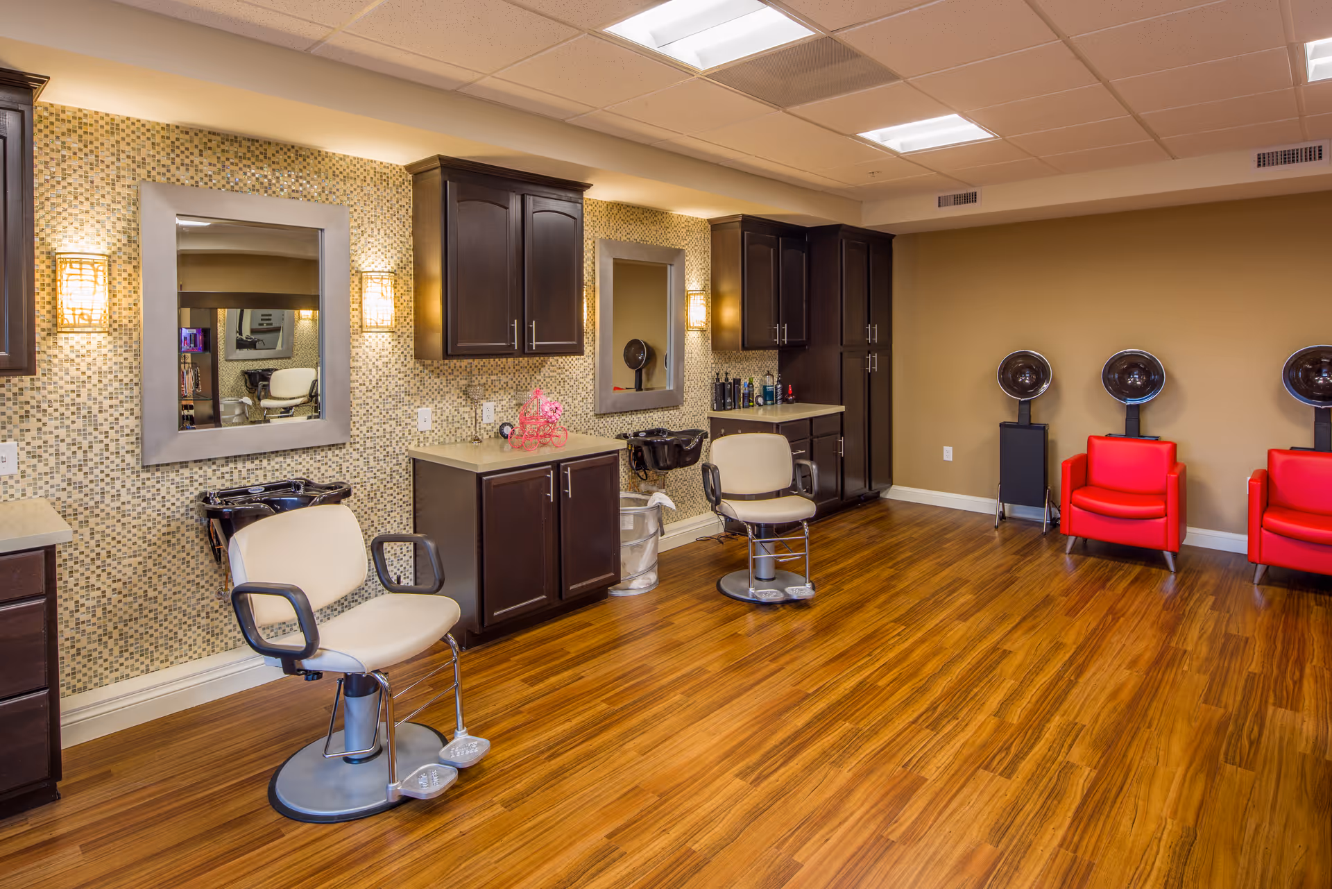 Interior of a hair salon area in an assisted living facility featuring two salon chairs in front of mirrors with dark wood cabinets above and below the countertops. The walls have a mosaic tile design behind the mirrors and a plain beige color elsewhere. On the right side, there are three red salon chairs with hair dryers positioned behind them. The floor is a polished wood finish.
