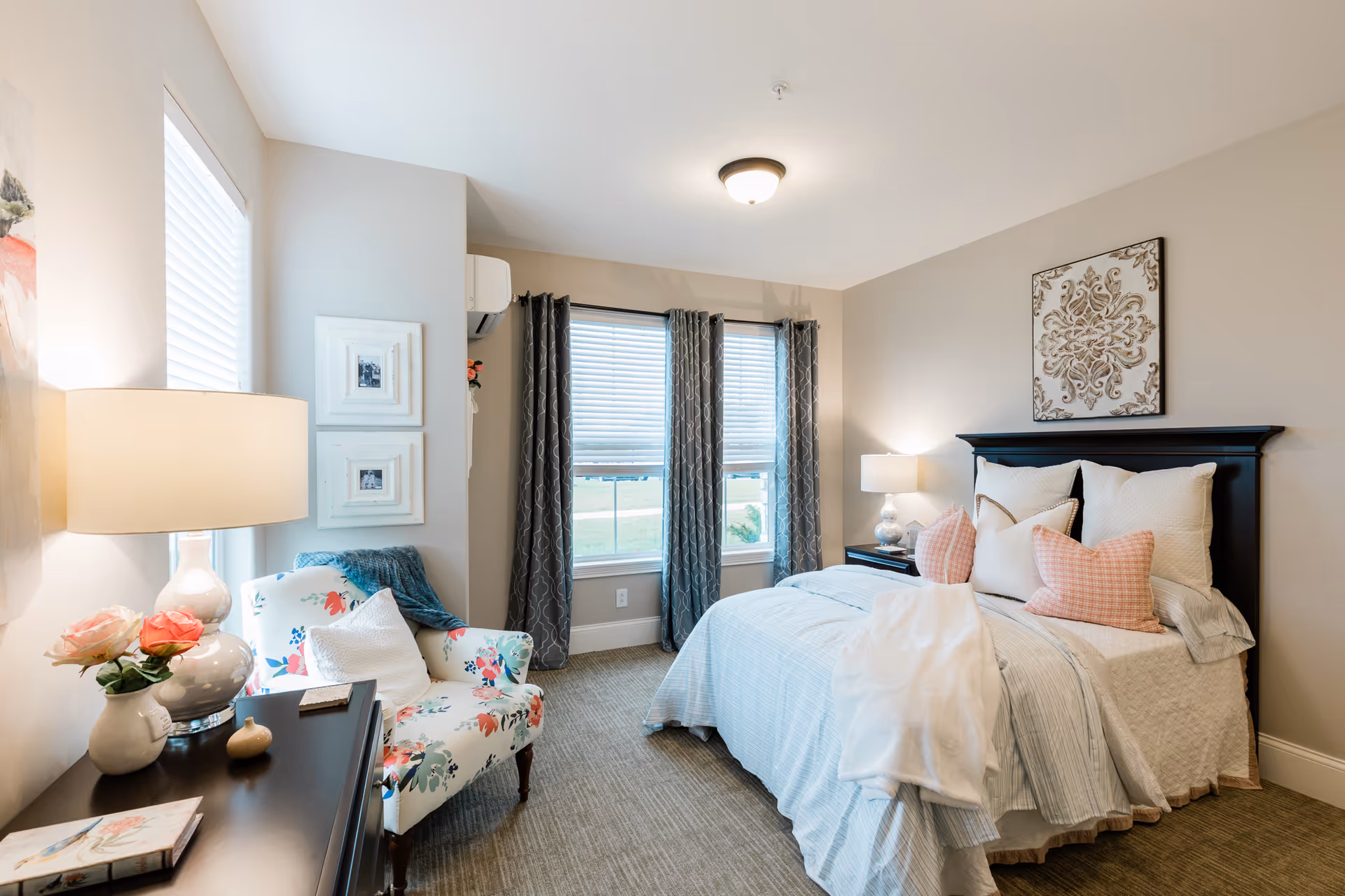 A bright and cozy bedroom with a neatly made bed featuring white and pink pillows and a white blanket. There is a black headboard and a decorative wall art above the bed. Next to the bed is a nightstand with a lamp. A floral patterned armchair with a white pillow and a blue throw blanket sits near a window with white blinds and gray curtains. A table lamp and a vase with pink flowers are on a dark wooden dresser. The room has beige walls and carpeted floor.