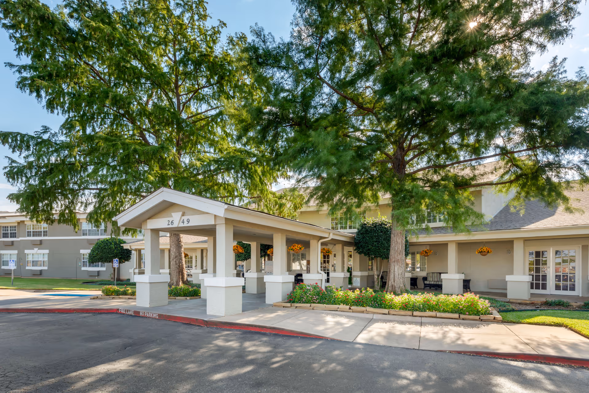 Entrance of Brookdale Sikes Lake facility with a covered drop-off area, surrounded by large trees and well-maintained landscaping including flower beds and trimmed bushes. The building is light-colored with multiple windows and a clear blue sky overhead.